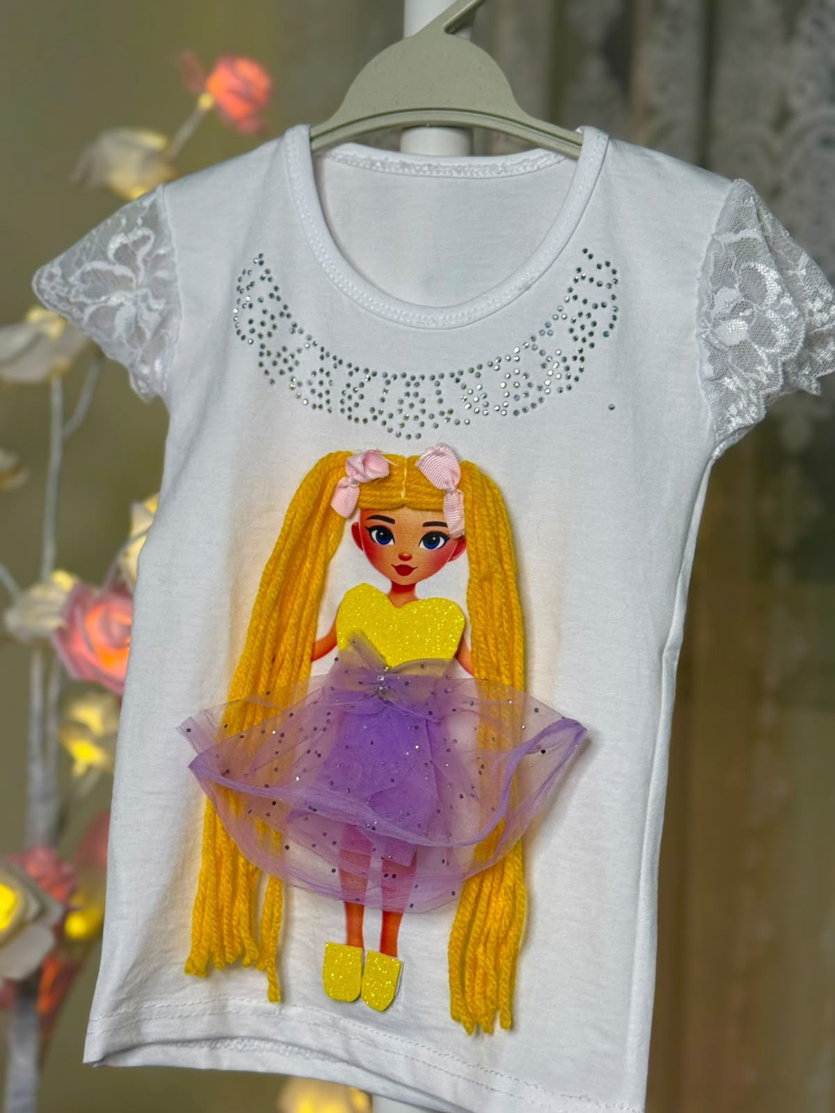 White lace-sleeve girls T-shirt on hanger with handmade doll appliqué featuring long yellow yarn pigtails with pink bows, yellow bodice, and purple sparkle tutu skirt