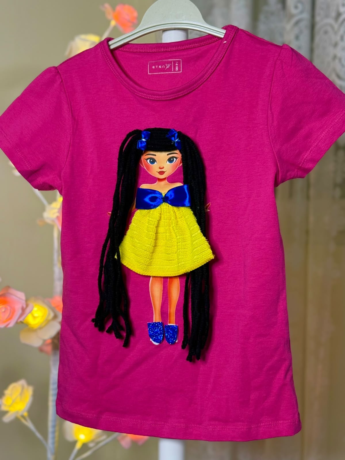 Pink girls T-shirt on hanger showing handmade black-haired doll appliqué in yellow dress with blue bow, photographed with decorative lights