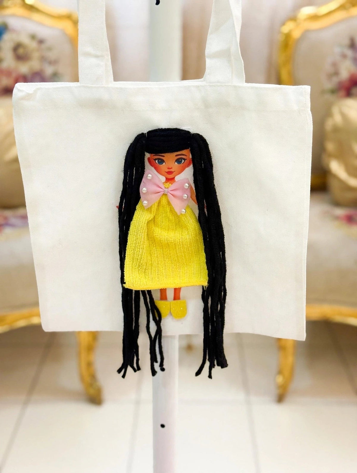 White canvas tote bag with handmade doll appliqué featuring long black yarn hair in pigtails, pink pearl bow, yellow textured dress, and yellow shoes