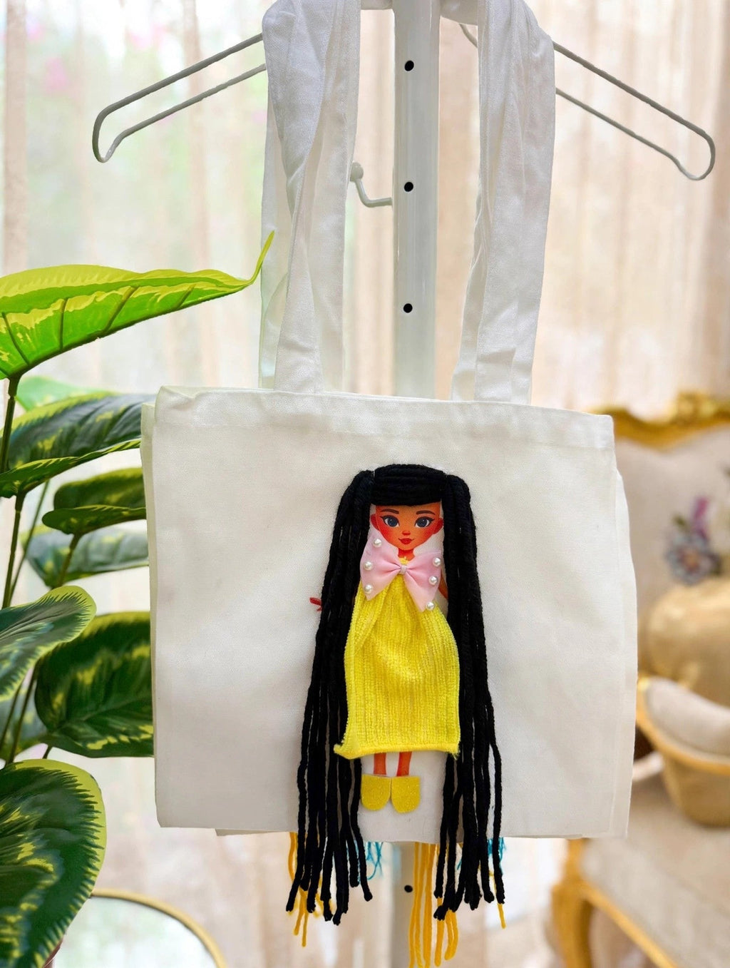 White canvas doll tote bag with black-haired doll in yellow dress hanging on a display stand beside green plants