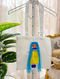 White canvas tote bag on display stand with handmade doll appliqué featuring long blue yarn hair, sparkly blue bow, yellow dress, and blue shoes