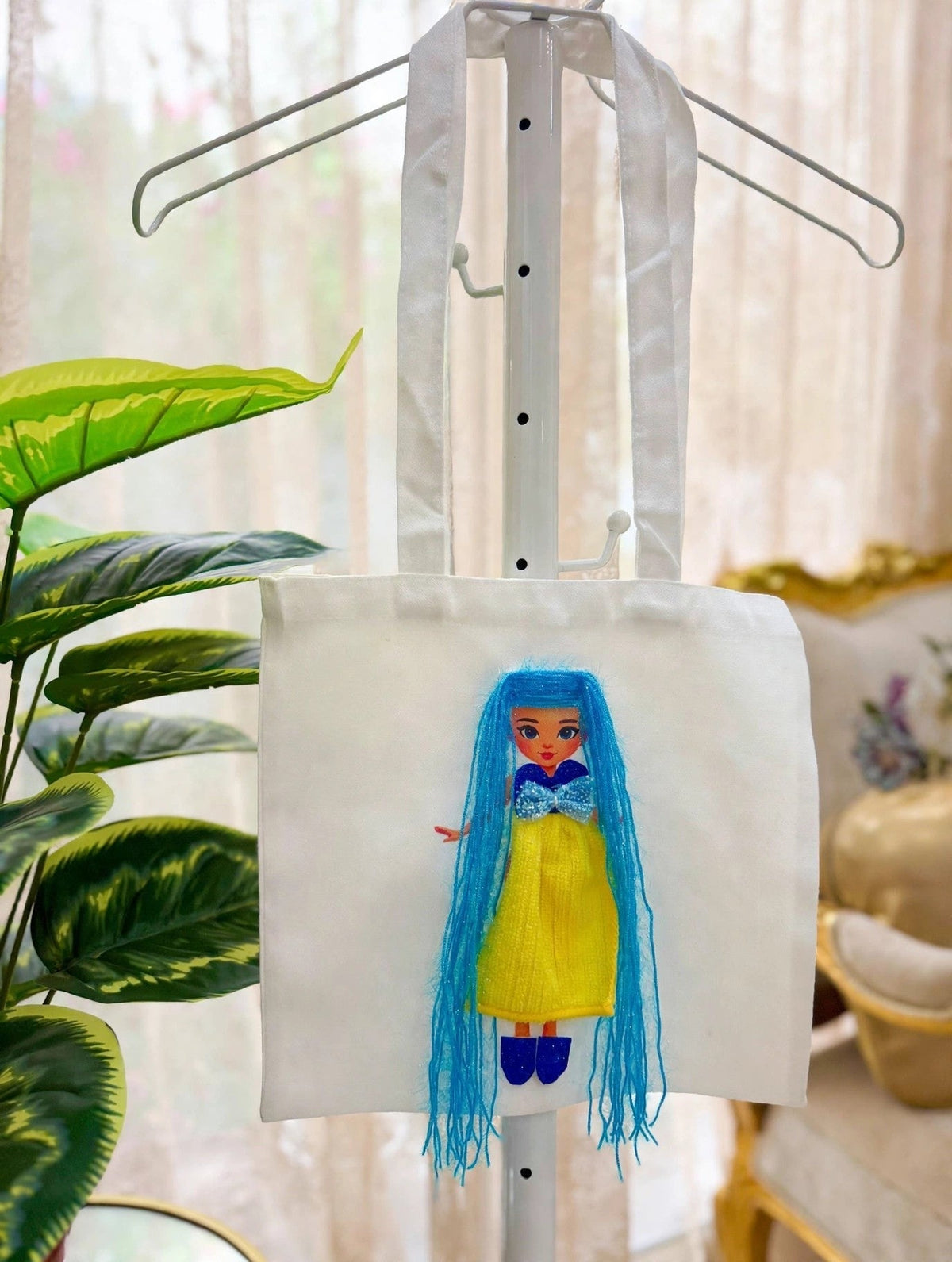 White canvas tote bag on display stand with handmade doll appliqué featuring long blue yarn hair, sparkly blue bow, yellow dress, and blue shoes
