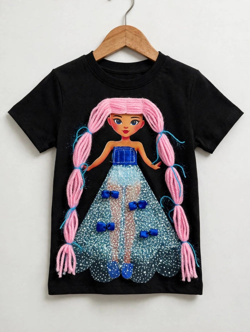 Styled photo of black girls T-shirt on pearl hanger with 3Sisters Fashion branding, featuring handmade doll appliqué with pink yarn pigtails and blue sparkle dress with bows
