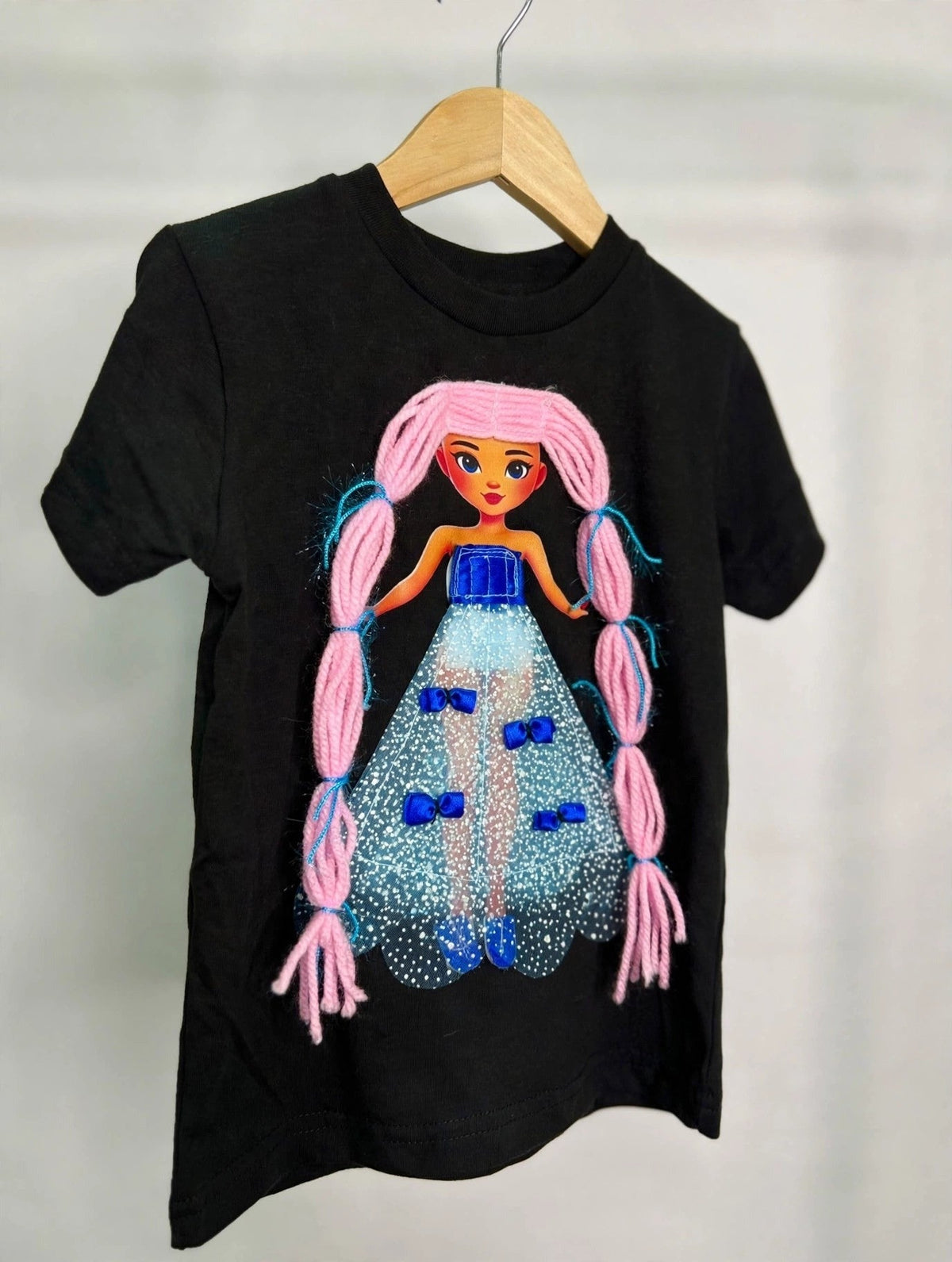 Black girls T-shirt on wooden hanger with handmade pink-haired doll appliqué in sparkly blue dress with satin bows