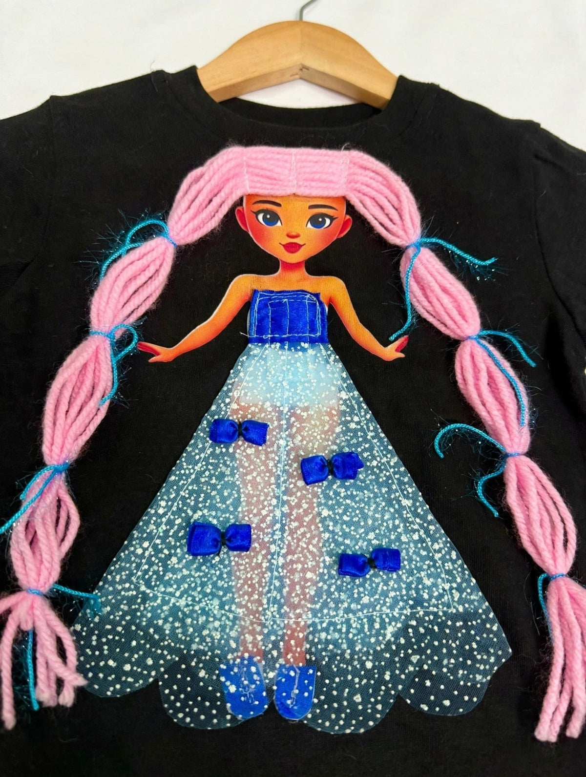 Close-up detail of handmade doll appliqué on black T-shirt showing pink yarn hair, blue bodice, and glittery tulle skirt with blue bows
